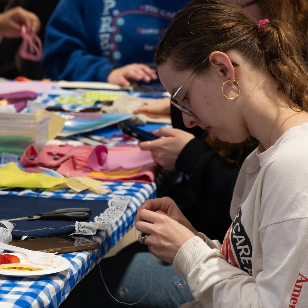 A photo of a student, in profile, working on their sewing project; fabrics, sewing supplies, and other materials are scattered in front of them on a table covered with a blue gingham tablecloth.