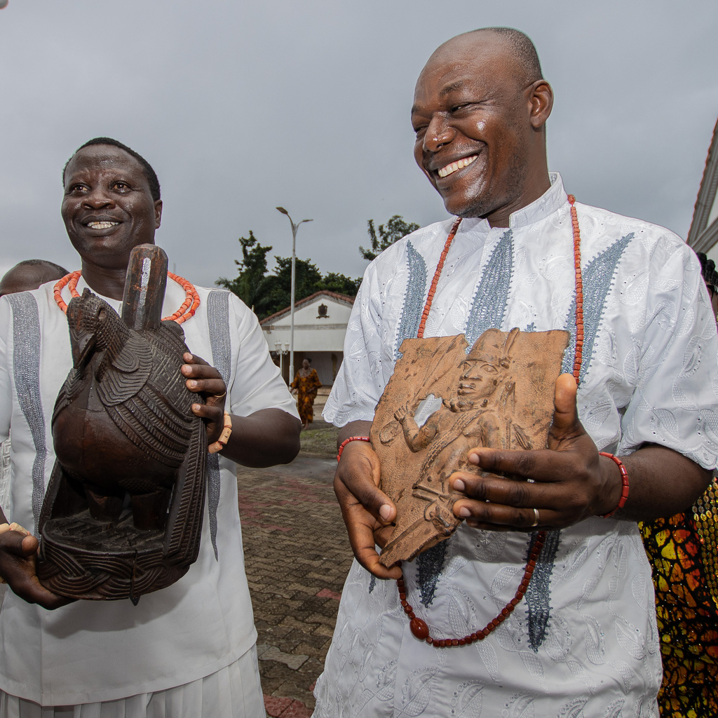 Two men wearing white, holding art objects and smiling