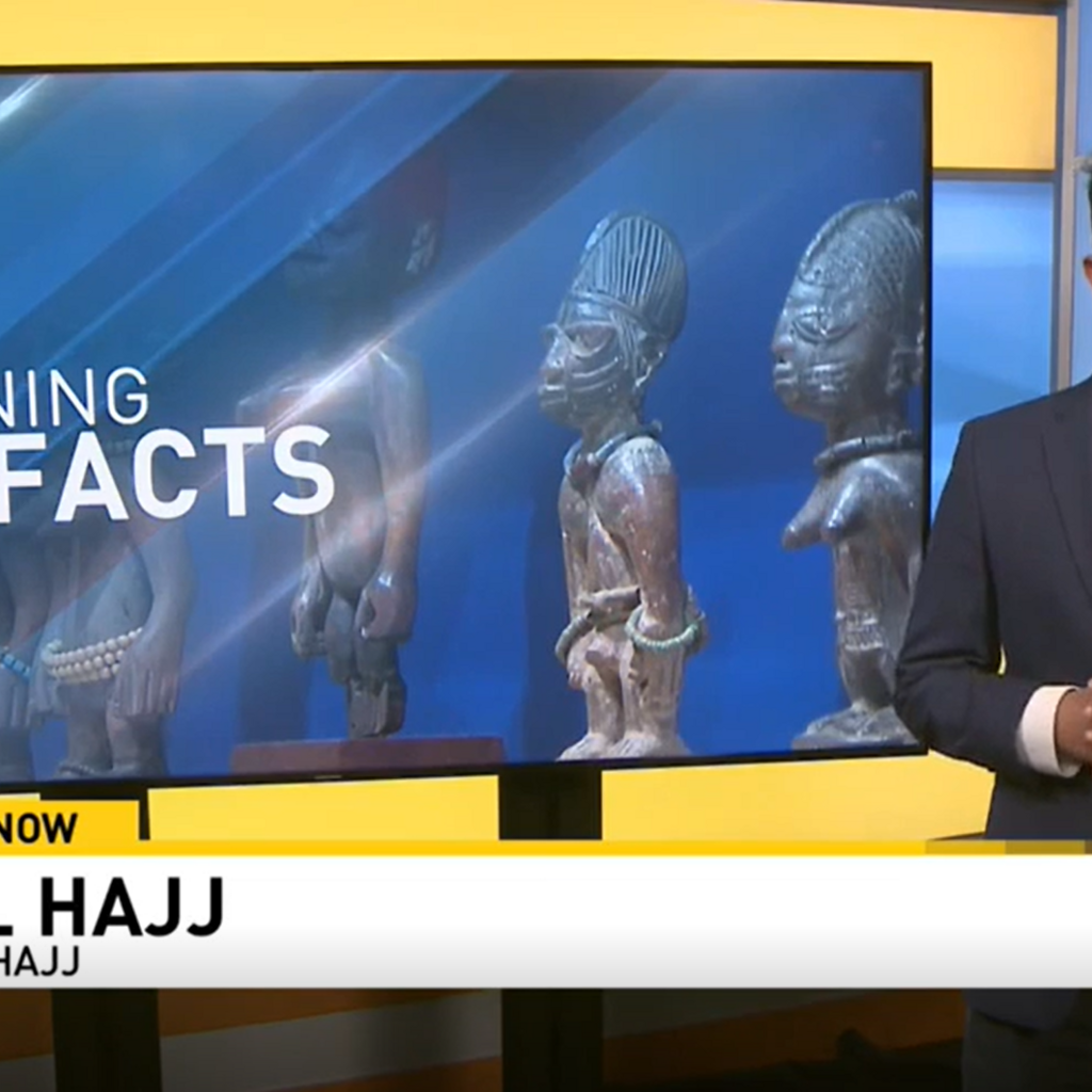 A person in a suit presenting the news standing next to a tv screen that reads "Returning Artifacts"