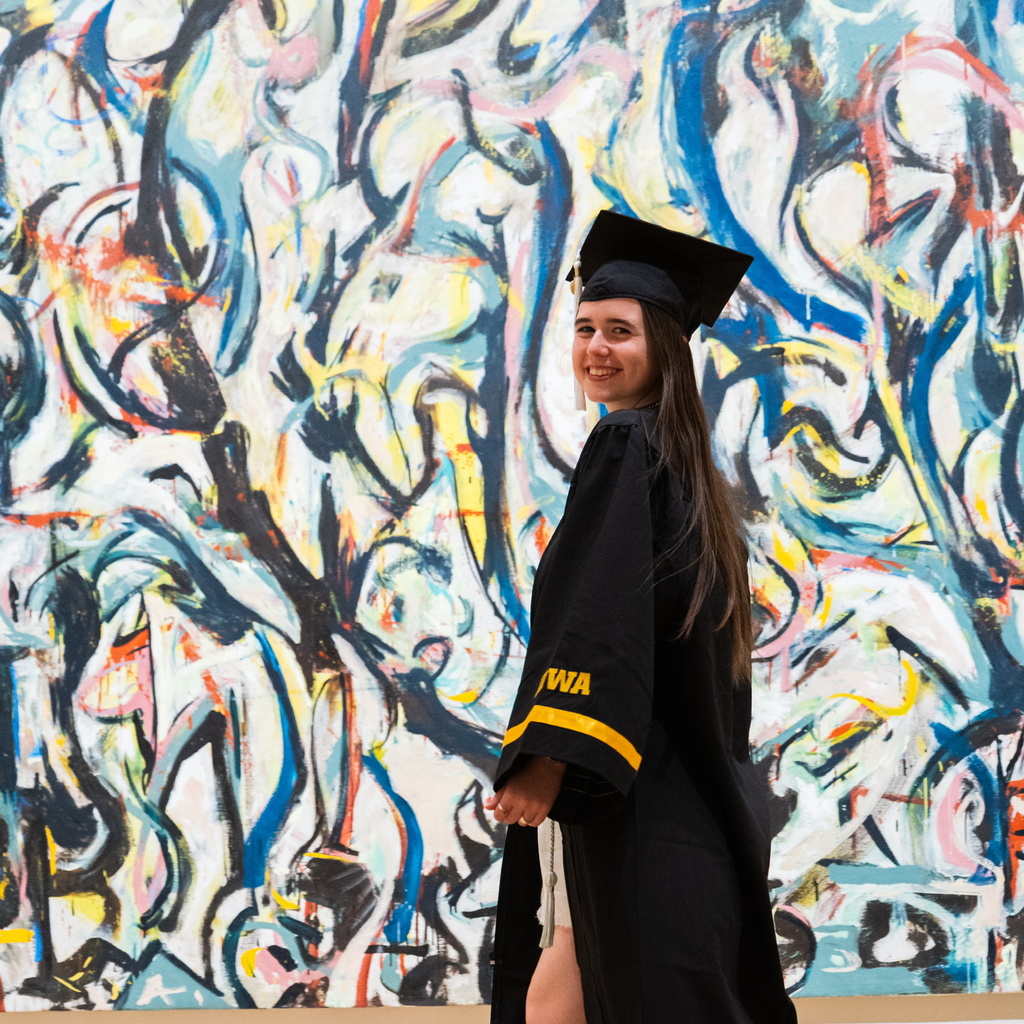 Gallery Host and Campus Engagement Coordinator Annelies Knight poses in her graduation gown in front of Jackson Pollock's Mural.