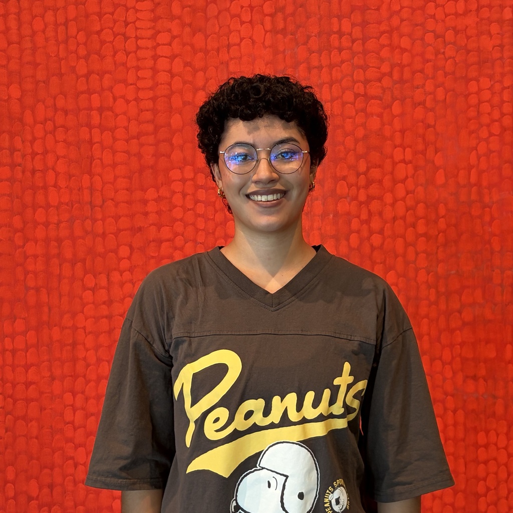Malenna poses, pictured from the waist up, in front of a red painting; she is smiling and wearing glasses.