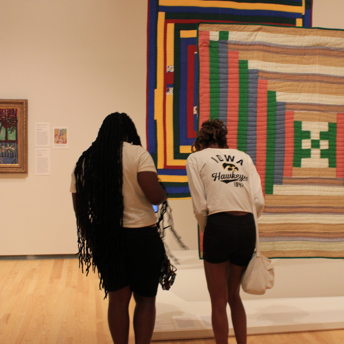 Two women with dark hair stand in front of brightly colored quilts hanging in a museum gallery