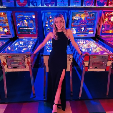 A photo of Faith McPhillips: she's wearing a black dress and standing amidst arcade games in a blue-lit room.
