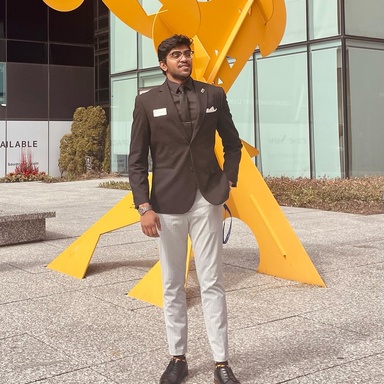 A photo of Hari Saravanan: he is posing in a suit in front of a yellow outdoor sculpture.