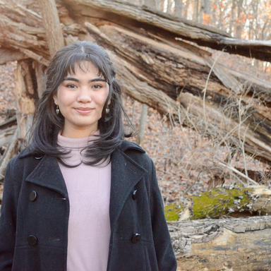 A photo of Jasmine Lee: she is standing in a black coat and a pink sweater in a forest.