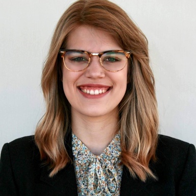 A photo of Maddie Meylor: she is against a white backdrop and dressed in a blazer, smiling at the camera.