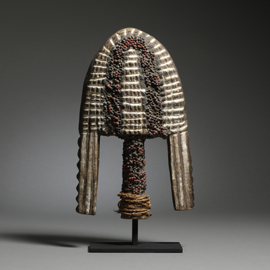 African crest mask with a tall, elongated shape, detailed with vertical lines, curved extensions, and red and black bead embellishments, mounted on a black stand against a dark background.