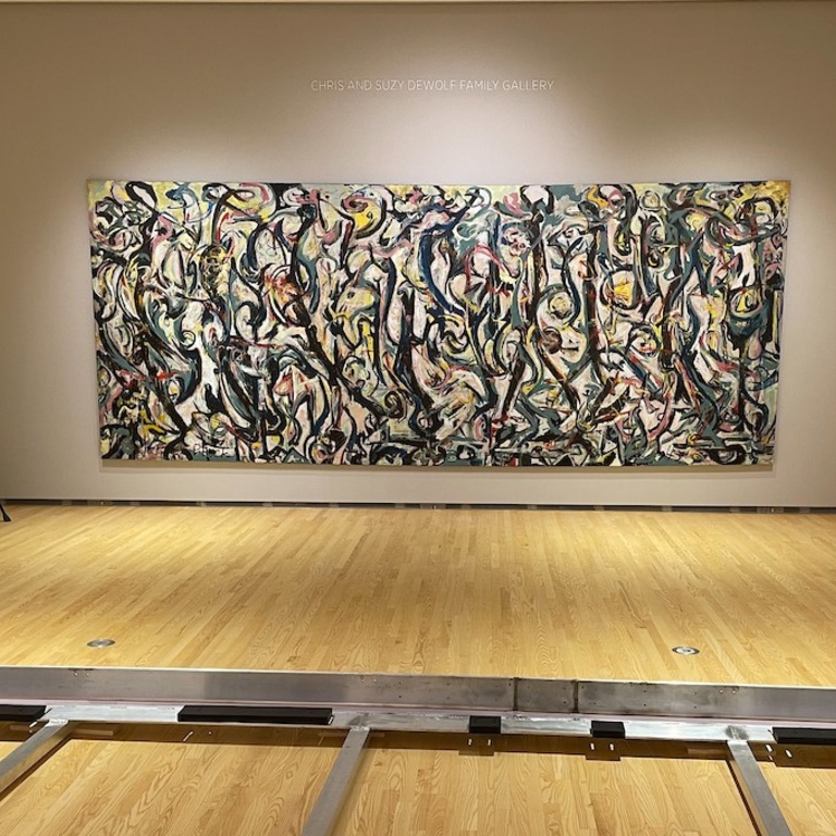 A large abstract painting with swirls of yellow, pink, teal, and light greenish blue, amid longer, vertical, curved black lines that have a quality of dance-like movement shown on a gallery wall with an aluminum travel frame on the floor in foreground.
