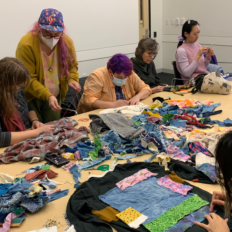 A shot of the table in the seminar room. it is covered in colorful bits of scrap fabric for people to use to patch their garments. Five people sit across the table, facing the camera. One person is in front of the camera, facing away, looking at the garment that they are mending.