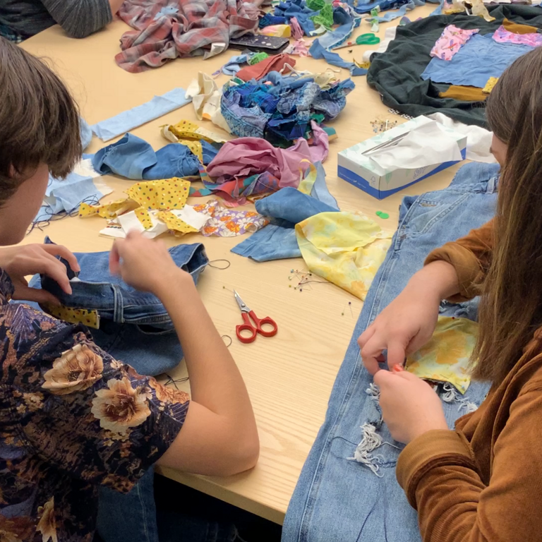 An image of two students sitting together, both bent over their work. The one on the left pulls thread through a garment that they are patching. The one on the right adjusts and makes stitches into the garment she is mending; a pair of ripped jeans with a yellow floral patch she's added.