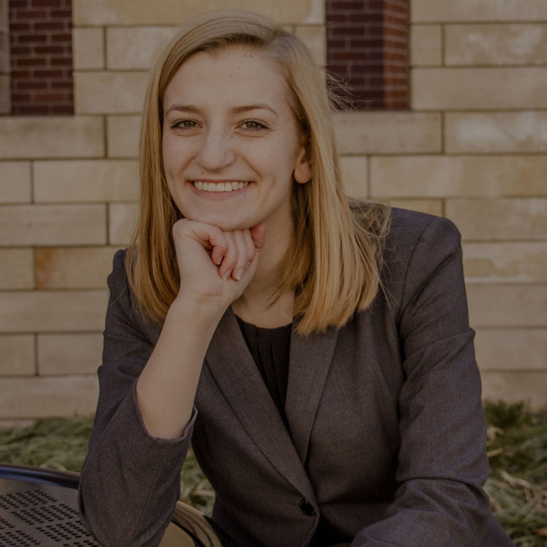 A photo of Eleanor Hildebrant: she is smiling at the camera, posting in front of a brick building, one hand propping up her chin.