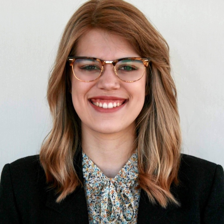 A photo of Maddie Meylor: she is against a white backdrop and dressed in a blazer, smiling at the camera.