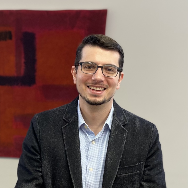 A photo of Nick Stroup: he is smiling at the camera, with an off-center red work of art hanging behind him.