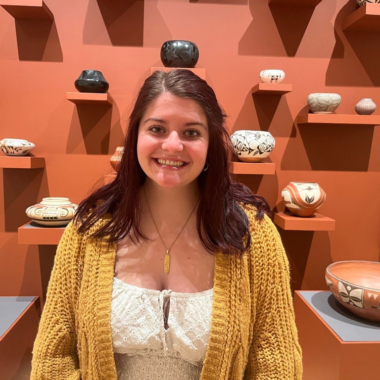 A photo of Alexis Belme posing in front of Native American ceramics.