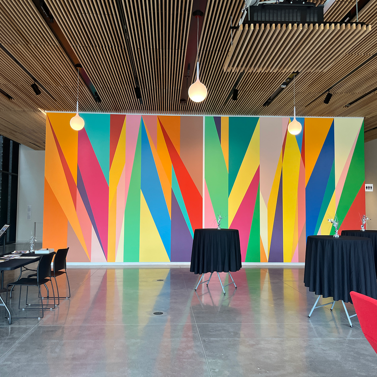 A photo showing the HBD, SMA set up. Facing west inside the building, black cocktail tables, an information desk with chairs and pamphlets, and the Stanley's rainbow-colored lobby mural can be seen.