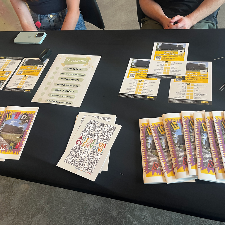 A photo of the HBD, SMA event information desk. On it are various paper goods--the zine specifically made for the event, and the Stanley Student Challenge stamp card.