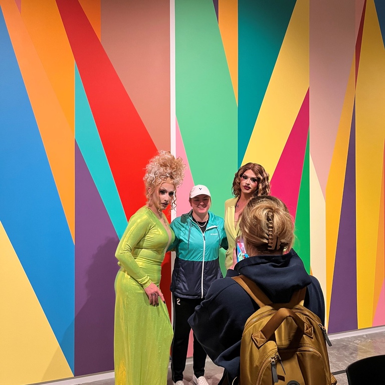 shown is a student standing in between two drag queens against the lobby mural 