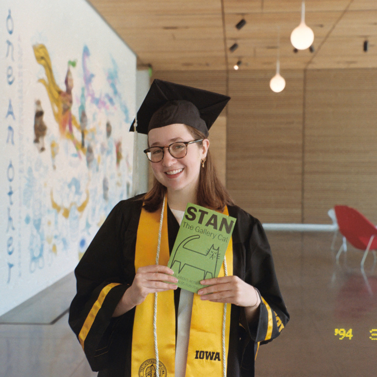 Museum Programs Intern Abbie McLaren, dressed in her graduation attire, poses with a Stan the Cat activity booklet, which she created in the spring of 2025, in the Stanley Museum of Art lobby.
