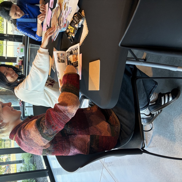 Three students sit around a table, actively collaging with piles of materials.