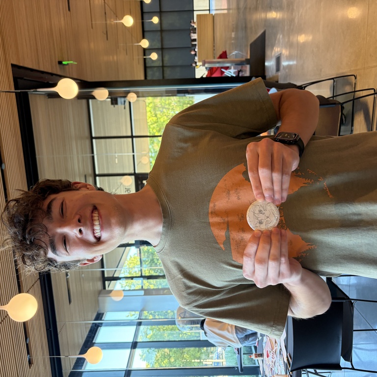 A college student poses, smiling, holding up a button they made in front of their chest.