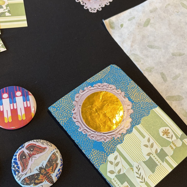 A flat lay photo of a small collaged notebook and collaged buttons with other collage materials on a black tablecloth.