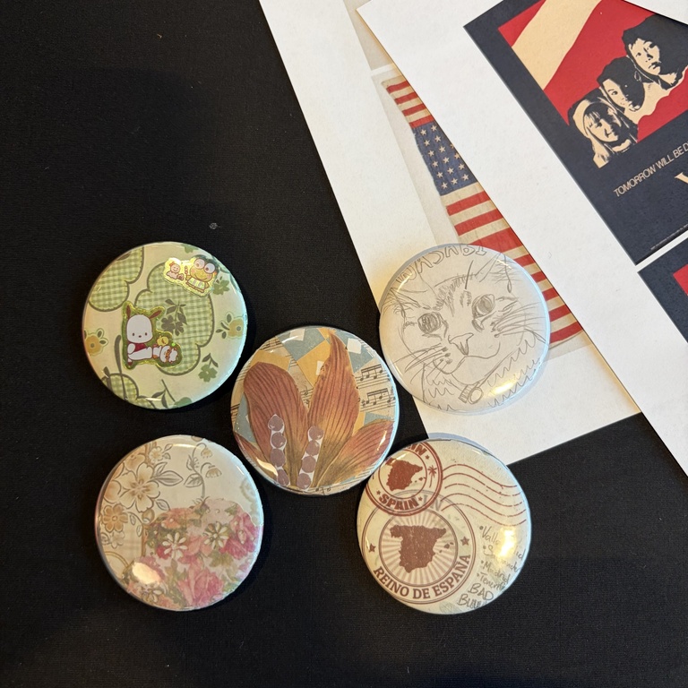 A flat lay photo of five collaged pinback buttons on top of a black tablecloth.