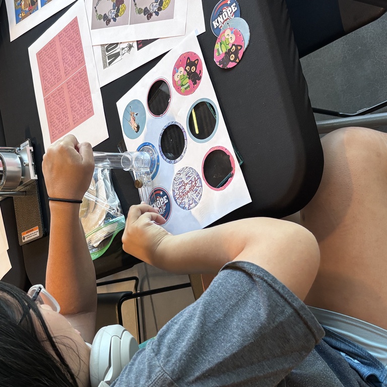 A student sits at a table, cutting out circular pieces of paper to use with the button maker.