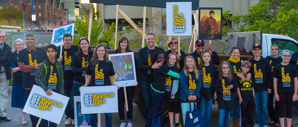 Group of people standing outside. They are wearing matching t-shirts and holding signs in preparation for the UI Homecoming parade.