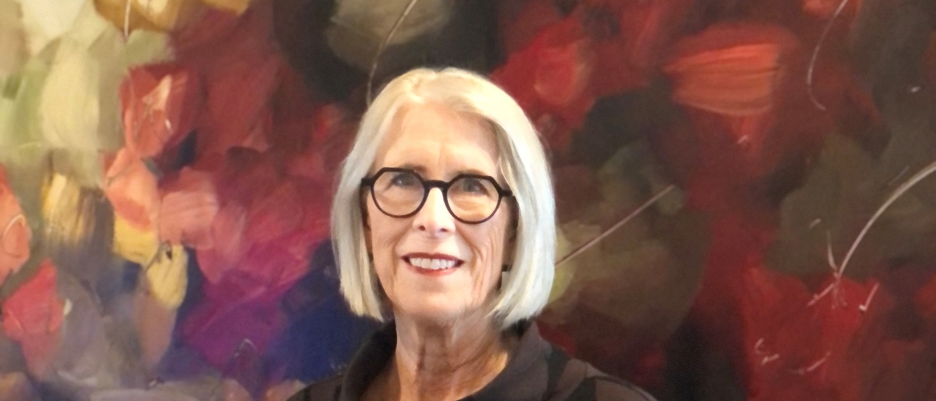 Woman with bobbed silver hair and dark-rimmed glasses wearing dark top with a gold brooch. She is standing in front of a painting.