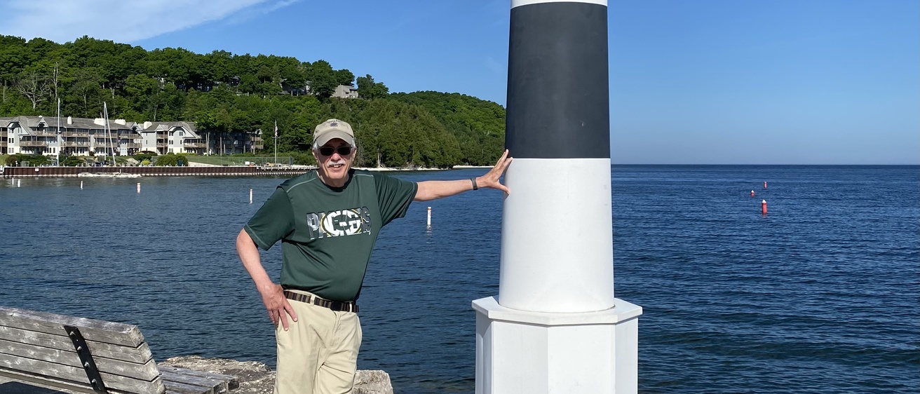 Man in blue shirt, khaki shorts and ball cap leaning against a small lighthouse