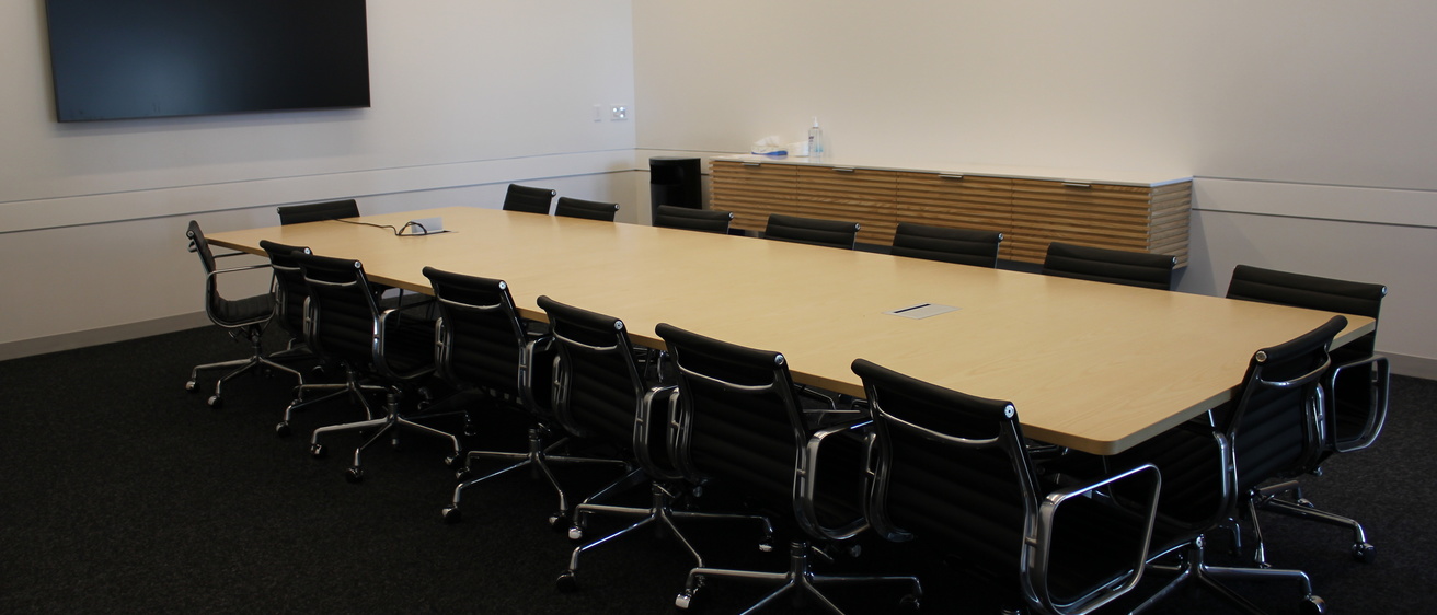 A meeting room with a long, wooden table surrounded by 16 black chairs. There is a large monitor mounted to the wall at left and a credenza along the far wall.