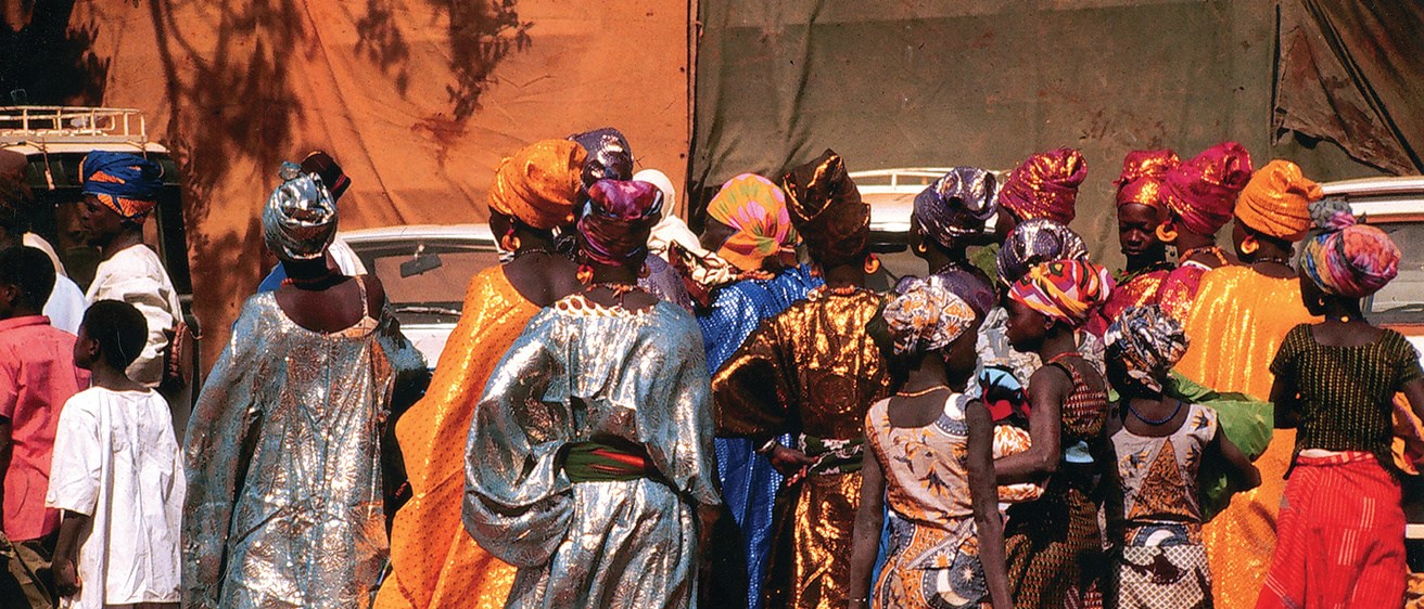 ​​​​​​​Fulani women attending the Ouahigouya regional fair. Colorfully dressed women with headwraps are seen from behind against an orange wall. 