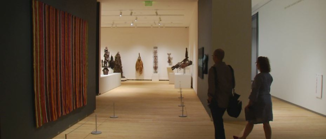 Two people walk through a museum gallery with textiles on the left and African masks in the background