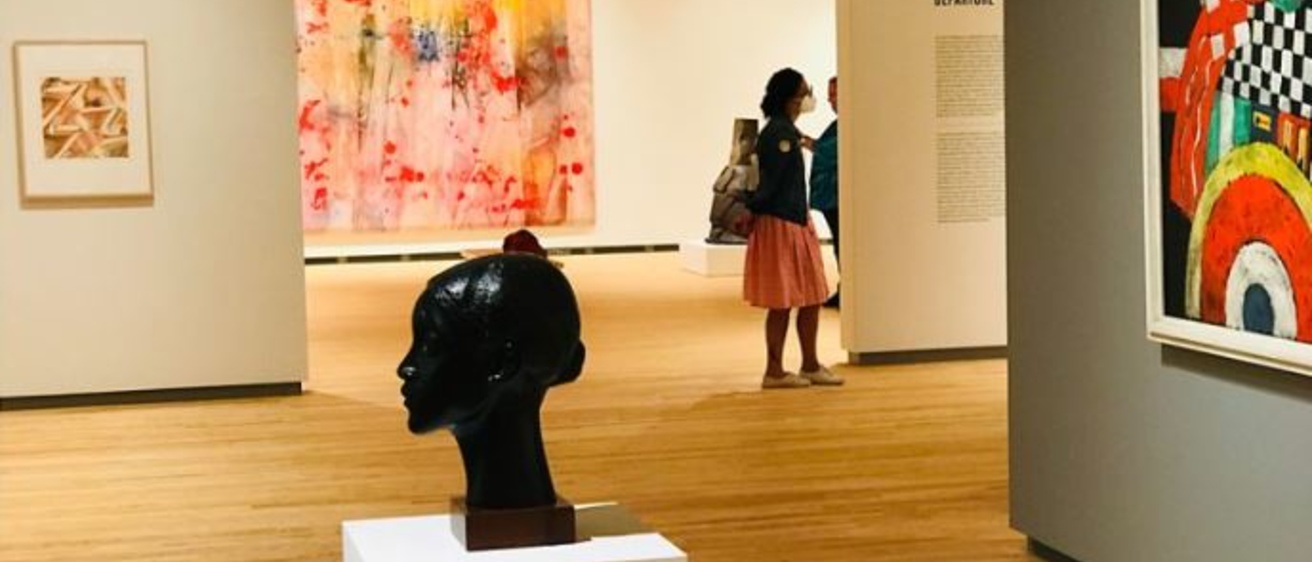An art gallery with a bronze bust in the foreground, a brightly colored, geometric painting on the right, and a person viewing a large multi-colored abstract painting in the background