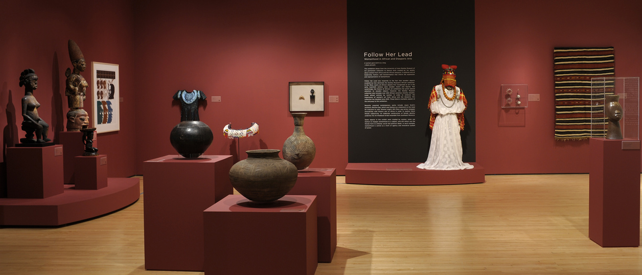 A museum gallery with dark red walls and light, wooden floor. There are large, African, ceramic pots in the foreground, and a mannequin with a white dress and jewelry in the background.