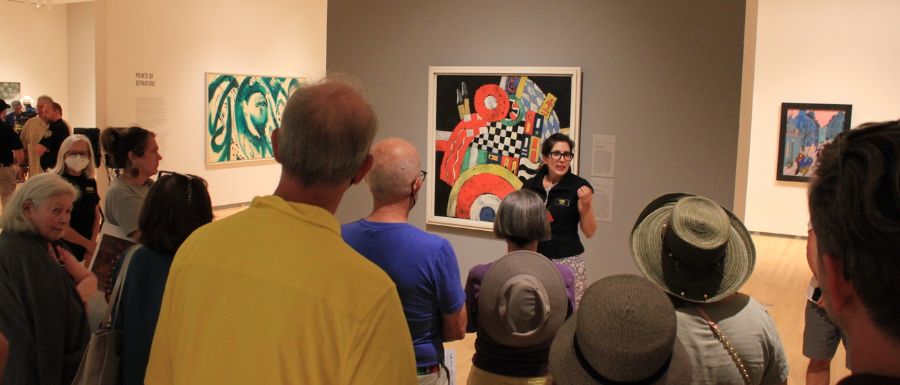 a woman in a black top and glasses stands in front of artwork in a gallery and speaks to a gathered group