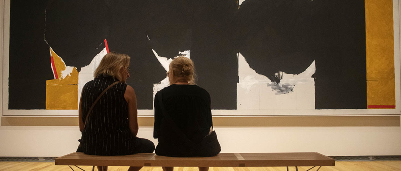 Two women sit on a wooden bench in front of a large wall painting
