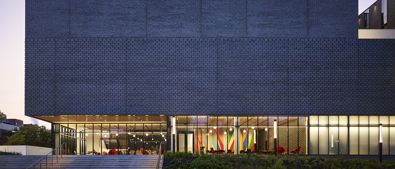 A grey, rectangular building is seen at dusk. The first level lobby is lit from within.