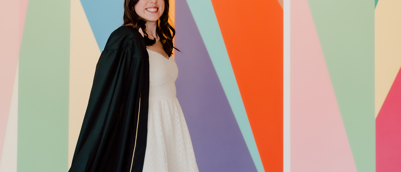 A photo of Anaka Sanders; it is an action shot of her striding in front of the lobby mural at the Stanley, dressed in her graduation robes.
