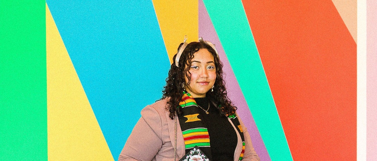 A photo of Lily Hester: they are standing in front of the mural in the Stanley's lobby, dressed up, posing with their graduation cap.