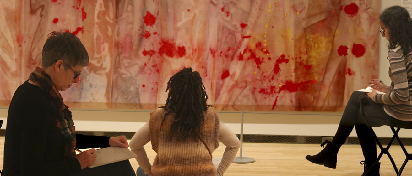 An Image featuring three people sitting in front of Sam Gilliam's "Red April," with notebooks.