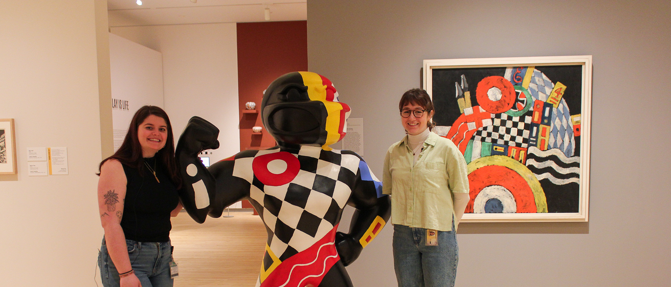 Two students stand in the galleries at the Stanley Museum of Art, posing next to a 6-foot-tall fiberglass Herky statue, that is painted to match a painting in the background of the photo--Marsden Hartley's "E."