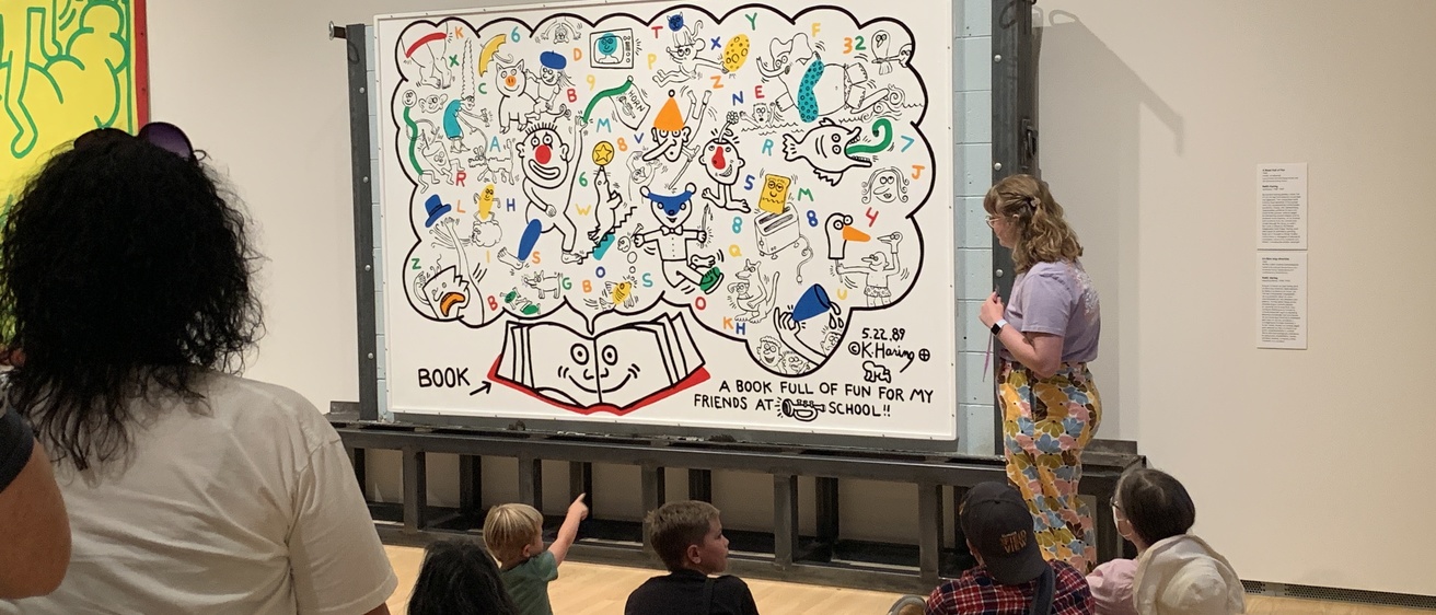 A photo of Bella Epstein, standing in front of the Keith Haring mural, "A Book Full of Fun," in the Stanley Museum of Art galleries, teaching in front of a group of children and their parents.
