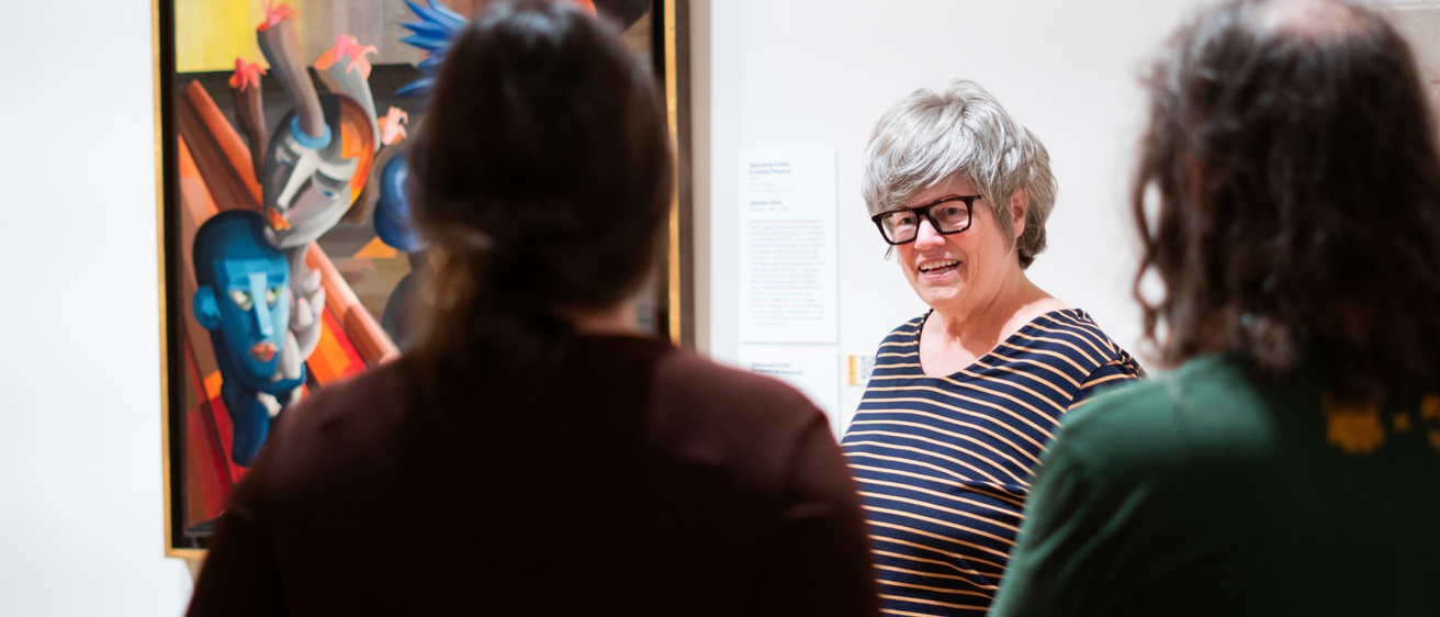 A person with glasses standing in front of a painting, speaking to a group
