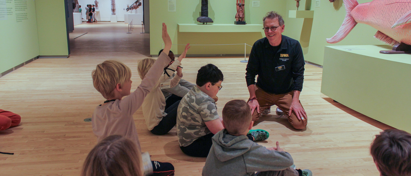 Children sitting on the floor of a gallery with an adult. One of the kids has his hand raised.