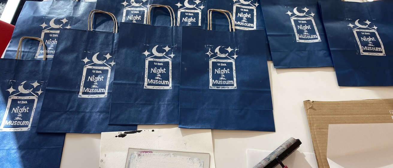 A photo of the entire setup: approximately one dozen printed blue paper bags are lined up along the top of a table, waiting to dry, set above a bottle of white ink, a plate with white ink on it, the ink covered linoleum block, an inked up brayer, and a clean brayer that has been used to apply pressure to the back of the plate to help transfer the ink.