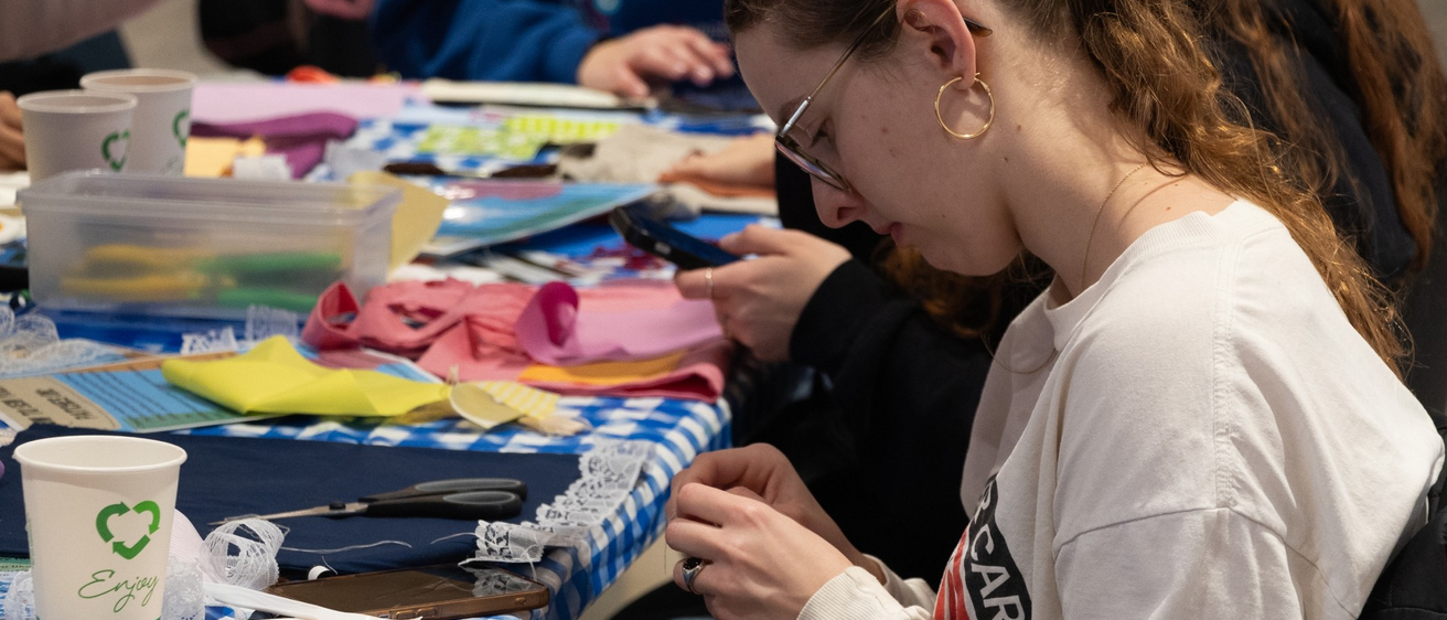 A photo of a student, in profile, working on their sewing project; fabrics, sewing supplies, and other materials are scattered in front of them on a table covered with a blue gingham tablecloth.
