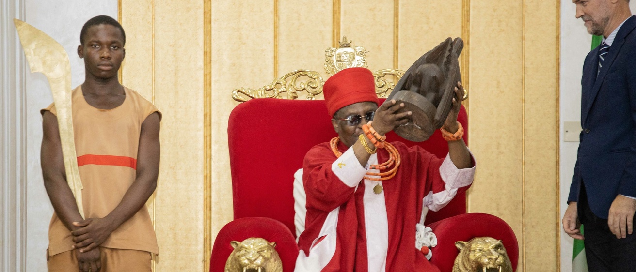 A man sitting on a red throne dressed in red robes holds up a wooden object