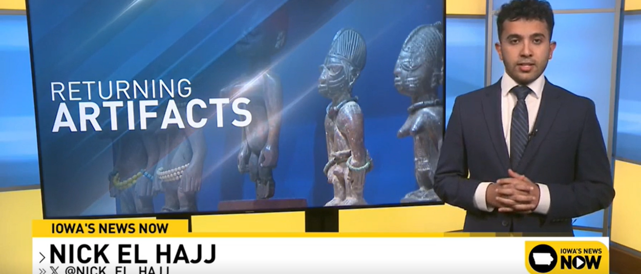 A person in a suit presenting the news standing next to a tv screen that reads "Returning Artifacts" 