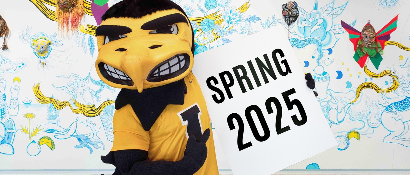 A black and yellow hawk mascot, Herky, holding up a sign that reads "Spring 2025"